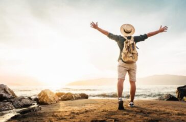 Happy man with backpack standing with arms up at the beach - Delightful tourist enjoying summer vacation by the seaside - Traveling life style and well being concept