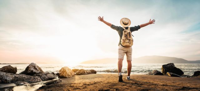Happy,Man,With,Backpack,Standing,With,Arms,Up,At,The Happy man with backpack standing with arms up at the beach - Delightful tourist enjoying summer vacation by the seaside - Traveling life style and well being concept
