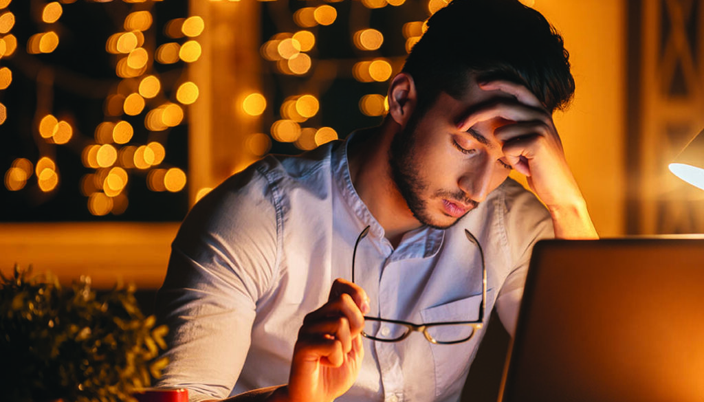 Feeling exhausted. Frustrated young man carrying eyeglasses and keeping eyes closed while sitting at his working place at night time with Christmas lights in the background