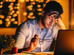 End-Of-Year Fatigue Feeling exhausted. Frustrated young man carrying eyeglasses and keeping eyes closed while sitting at his working place at night time with Christmas lights in the background