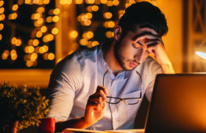 End-Of-Year Fatigue Feeling exhausted. Frustrated young man carrying eyeglasses and keeping eyes closed while sitting at his working place at night time with Christmas lights in the background