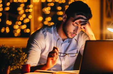 Feeling exhausted. Frustrated young man carrying eyeglasses and keeping eyes closed while sitting at his working place at night time with Christmas lights in the background