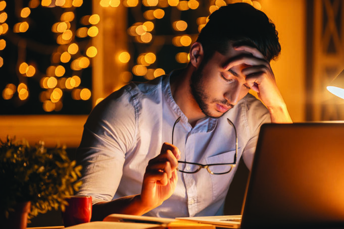 End-Of-Year Fatigue Feeling exhausted. Frustrated young man carrying eyeglasses and keeping eyes closed while sitting at his working place at night time with Christmas lights in the background