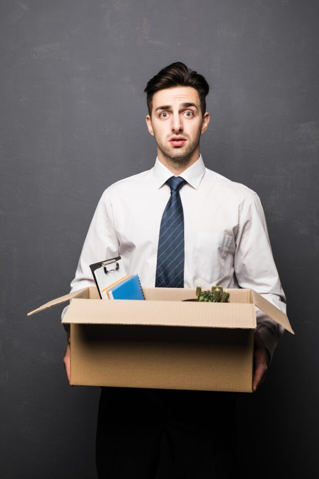 Getting,Fired.,Side,View,Of,Handsome,Businessman,In,Formal,Wear Getting fired. Side view of handsome businessman in formal wear holding a box with his stuff, on gray